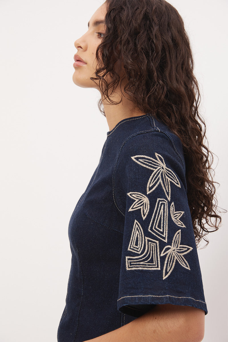 Woman wearing a stretch denim mini dress with a soft A-line silhouette, jewel neckline, short flared sleeves with contrast embroidery, and heavy contrast topstitching.