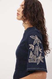 Woman wearing a stretch denim mini dress with a soft A-line silhouette, jewel neckline, short flared sleeves with contrast embroidery, and heavy contrast topstitching.
