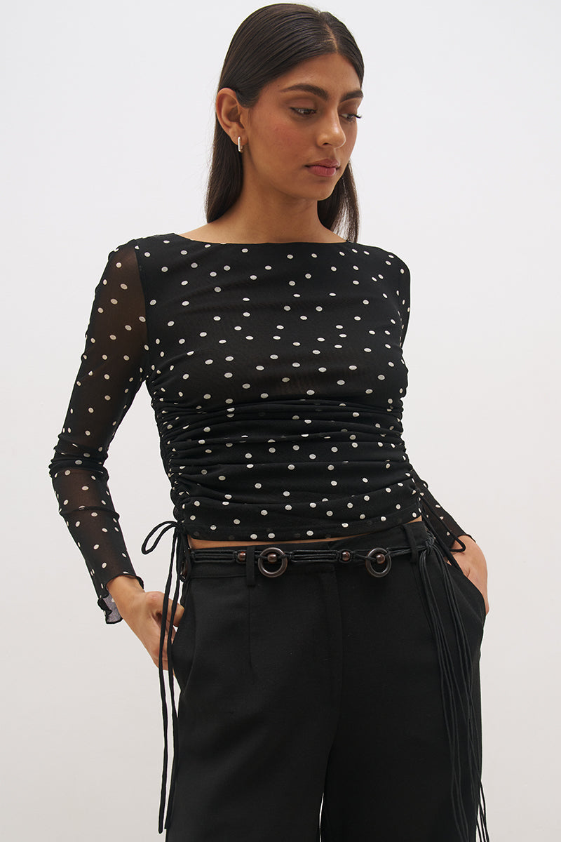 Woman wearing a printed polka dot mesh long sleeve longline top with a boat neckline, sheer sleeves, lettuce hem cuffs, and a drawcord waist detail.