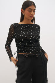 Woman wearing a printed polka dot mesh long sleeve longline top with a boat neckline, sheer sleeves, lettuce hem cuffs, and a drawcord waist detail.