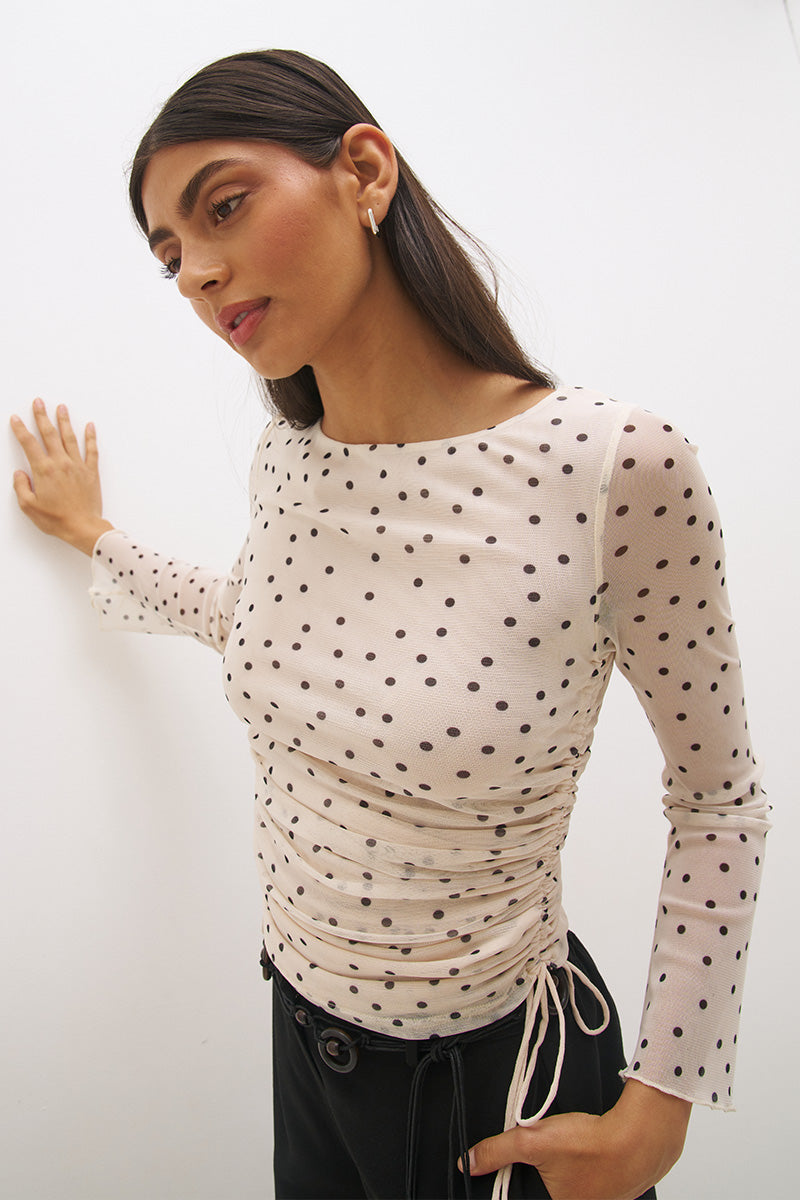 Woman wearing a printed polka dot mesh long sleeve longline top with a boat neckline, sheer sleeves, lettuce hem cuffs, and a drawcord waist detail.