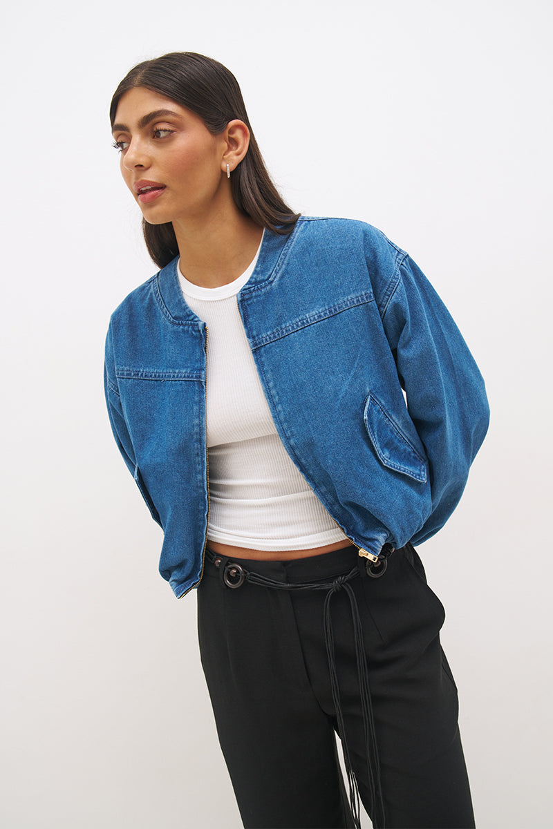 Woman wearing a washed denim bomber jacket with a stand collar, wide dropped shoulders, long balloon sleeves, bubble hem, and metal zip closure.