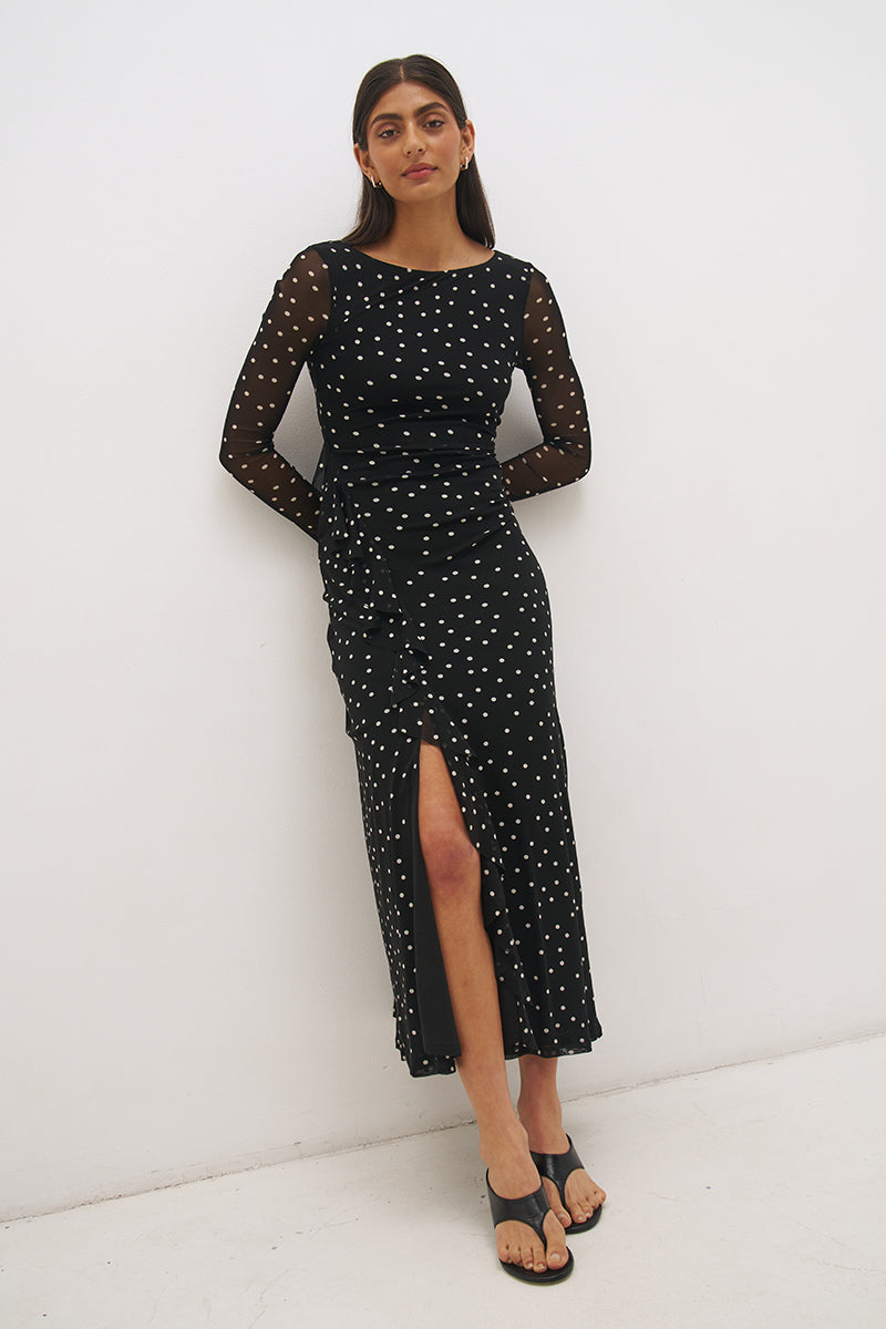 Woman wearing a printed polka dot mesh long sleeve midi dress in a column silhouette, with a boat neckline, sheer sleeves, bodice ruching, and a curved side frill split detail.