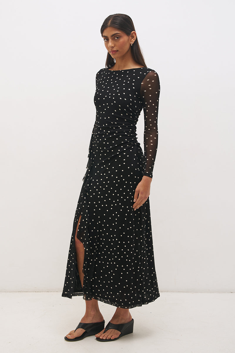 Woman wearing a printed polka dot mesh long sleeve midi dress in a column silhouette, with a boat neckline, sheer sleeves, bodice ruching, and a curved side frill split detail.