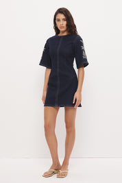 Woman wearing a stretch denim mini dress with a soft A-line silhouette, jewel neckline, short flared sleeves with contrast embroidery, and heavy contrast topstitching.