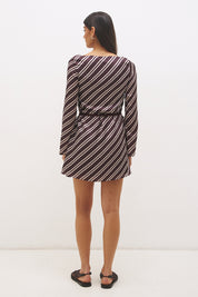 Woman wearing an asymmetric stripe satin mini dress with boat neckline, flared sleeves, flared hem, and removable waist tie