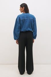 Woman wearing a washed denim bomber jacket with a stand collar, wide dropped shoulders, long balloon sleeves, bubble hem, and metal zip closure.