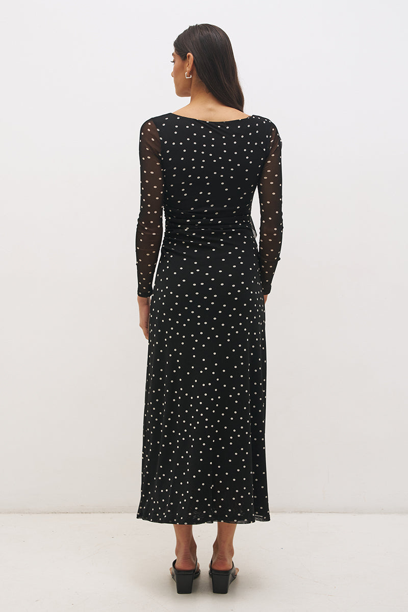 Woman wearing a printed polka dot mesh long sleeve midi dress in a column silhouette, with a boat neckline, sheer sleeves, bodice ruching, and a curved side frill split detail.