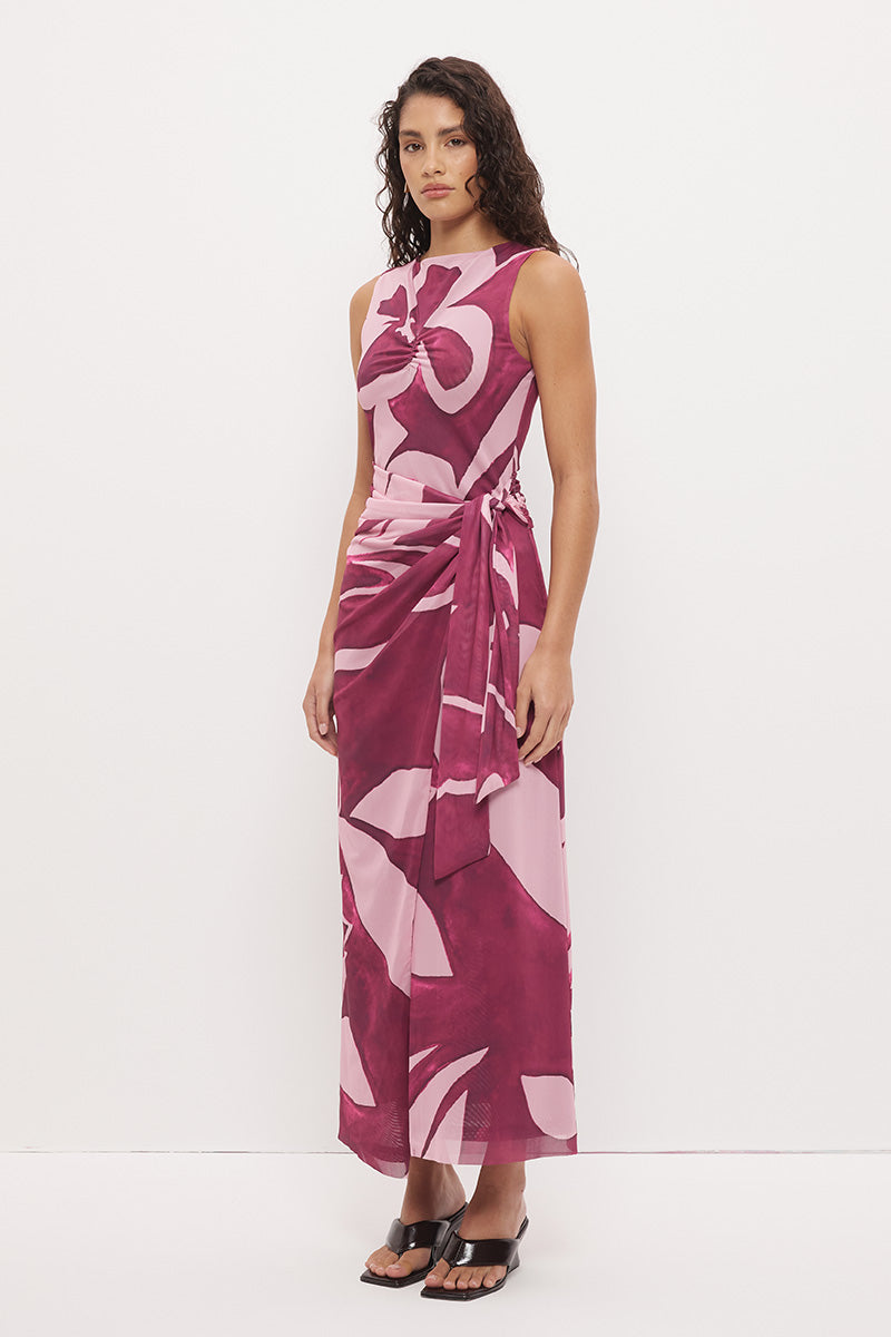 Woman wearing a sleeveless maxi dress with an exclusive placement print, sarong-style wrap skirt overlay, ruched asymmetric waistband, and boat neckline in a softly A-lined silhouette
