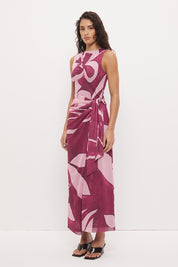 Woman wearing a sleeveless maxi dress with an exclusive placement print, sarong-style wrap skirt overlay, ruched asymmetric waistband, and boat neckline in a softly A-lined silhouette