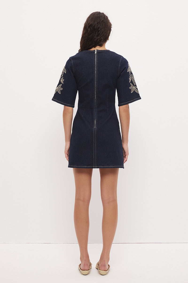 Woman wearing a stretch denim mini dress with a soft A-line silhouette, jewel neckline, short flared sleeves with contrast embroidery, and heavy contrast topstitching.